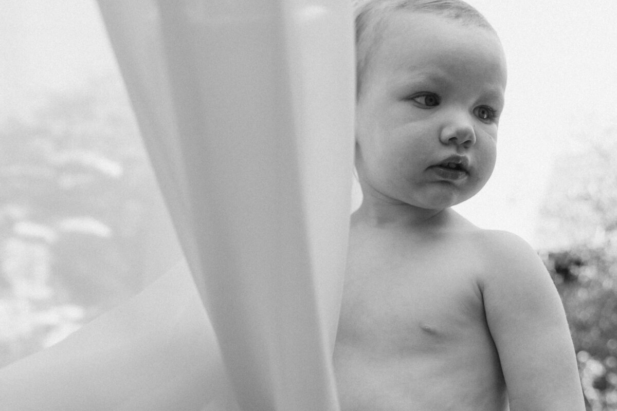 Black and white image of toddler girl standing next to sheer fabric, looking away