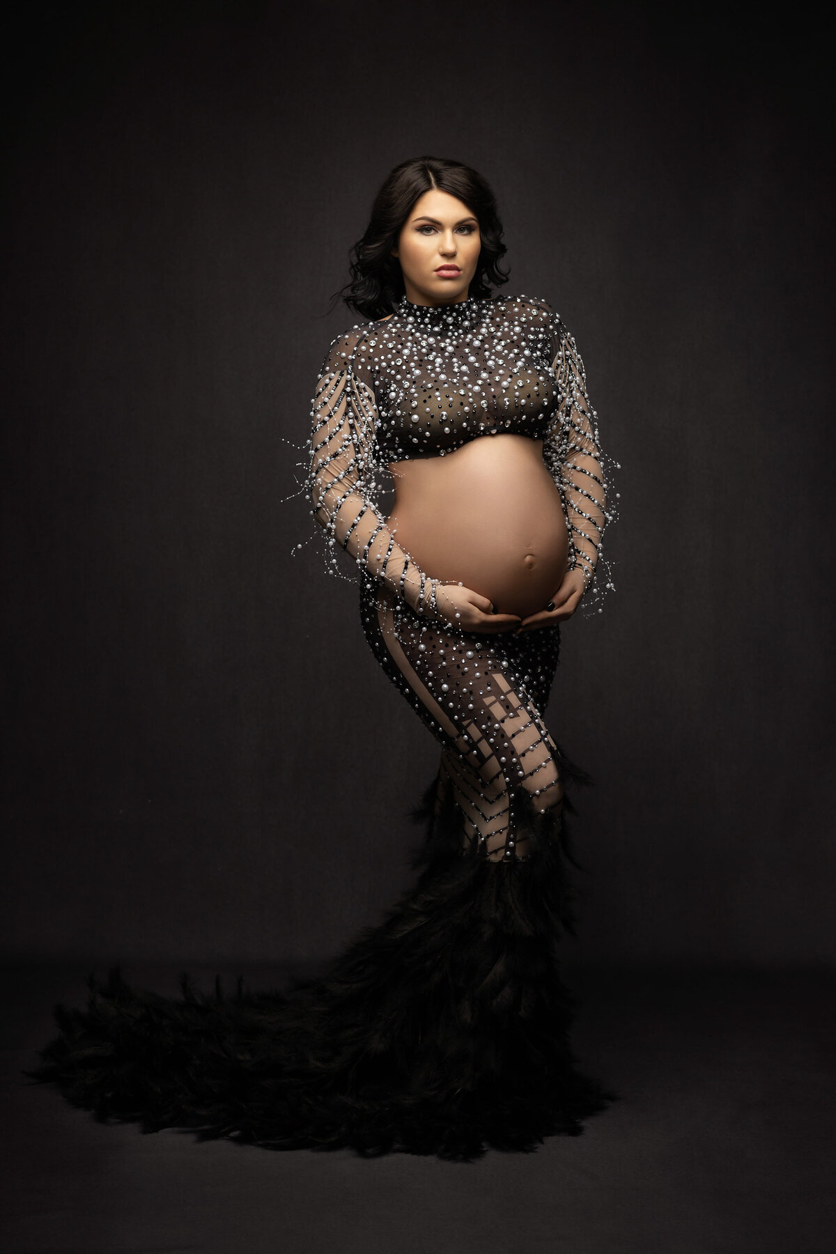  A captivating portrait showcasing a woman wearing a stunning black gown adorned with sparkling crystals and feathers, her graceful pose highlighted against a dark studio backdrop. Captured by Lindsay Walden Photography, this fine art portrait embodies luxury and elegance.