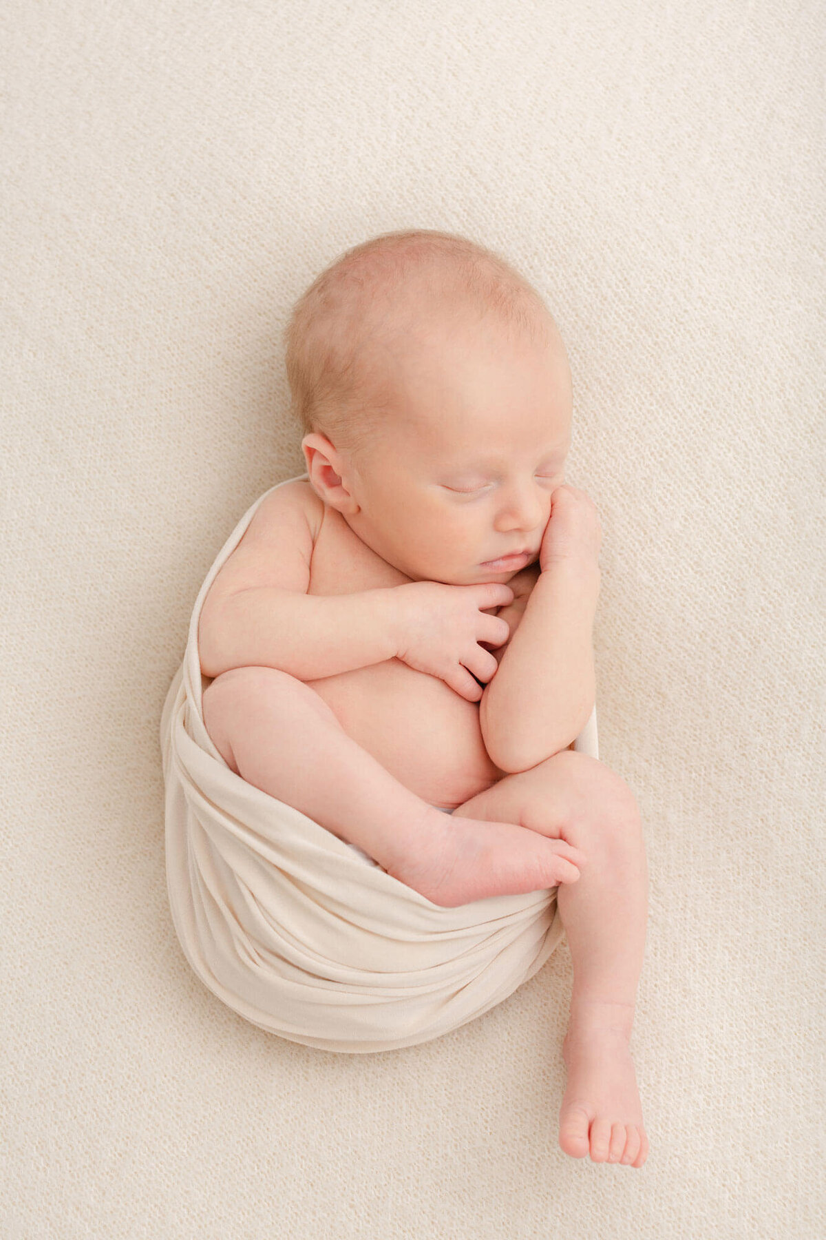 Baby swaddled in beige with arms and legs hanging out of swaddle, he looks peaceful and cozy and is laying on a beige blanket.