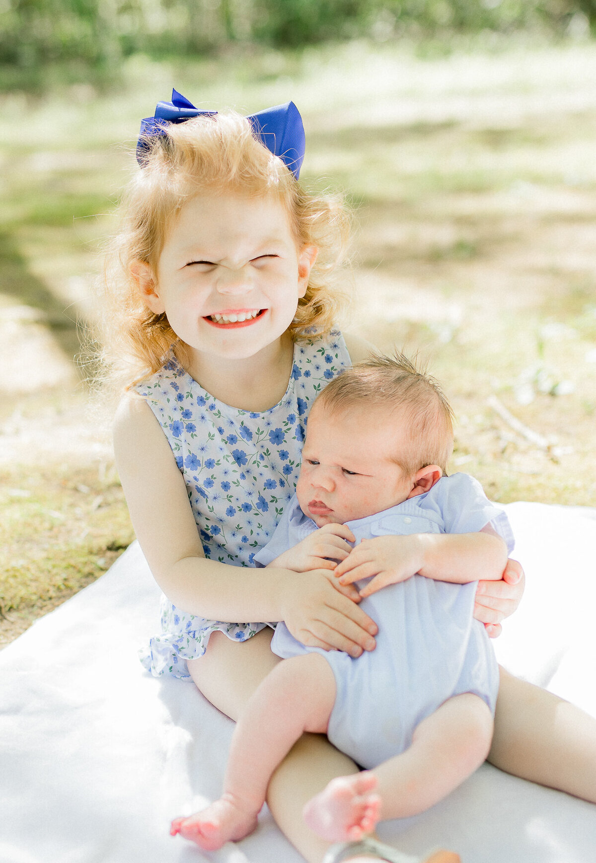 A proud big sister with curly red hair and a bright blue bow grins while holding her newborn baby sibling in a soft, natural outdoor setting. This sweet sibling moment captures the joy and tenderness of welcoming a new baby, reflecting the light and heartfelt style of a Birmingham newborn photographer.
