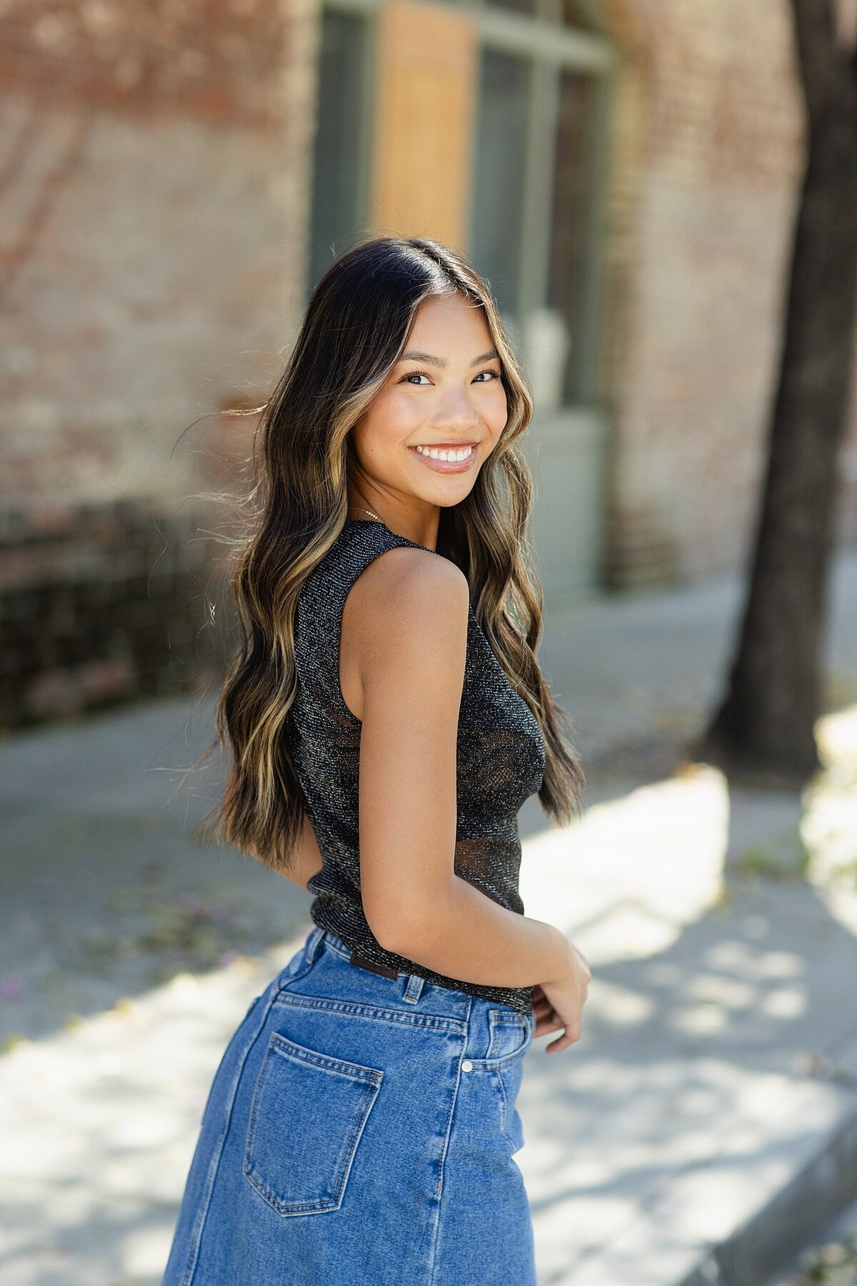 Smiling senior girl with long balayage hair and sleeveless top posing in natural light for an editorial high school senior photo in downtown Pasadena, California