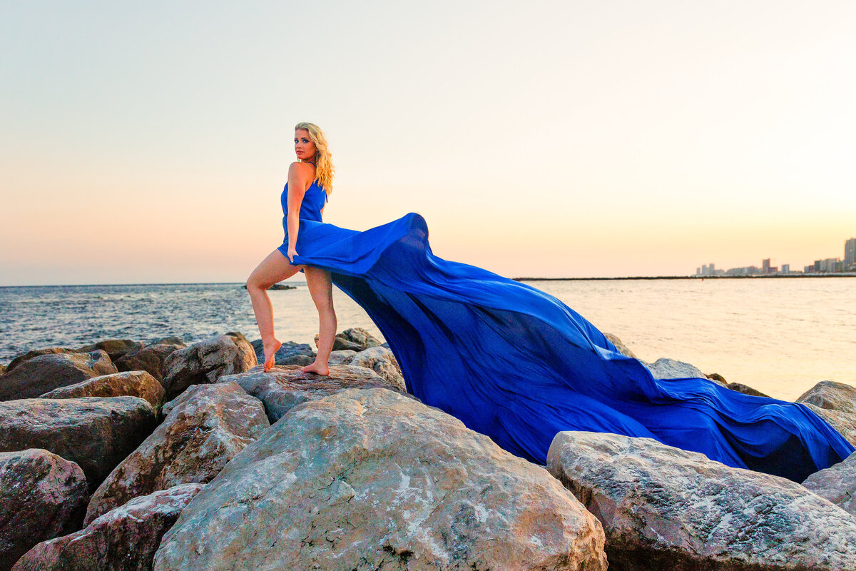 Girl looks over her shoulder while standing on seaside rocks.  The wind captures the 15 foot flying dress and flies in the wind.