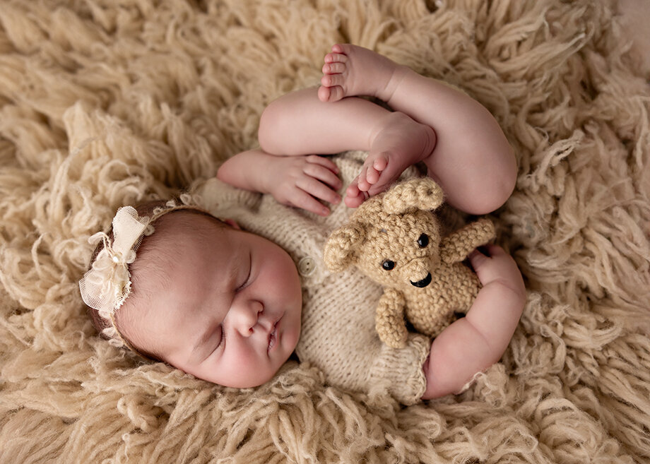 Baby girl in a beige romper.  Huck Finn pose with a boho headband.  She is holding a small teddy bear and laying on a beige flokati