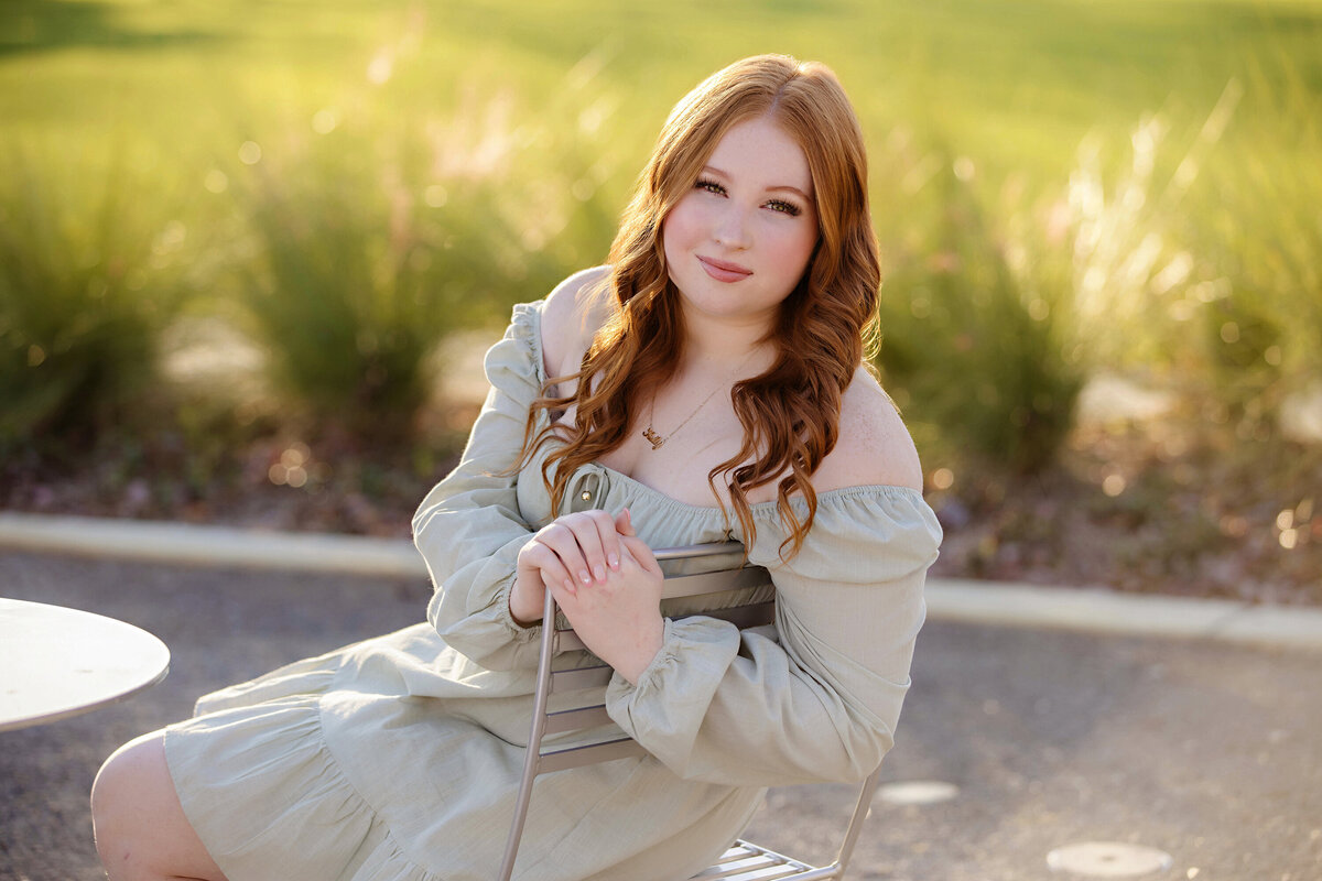 High school senior seated in metal chair with sage dress and tall grass behind her, framed by warm tones and gentle breeze