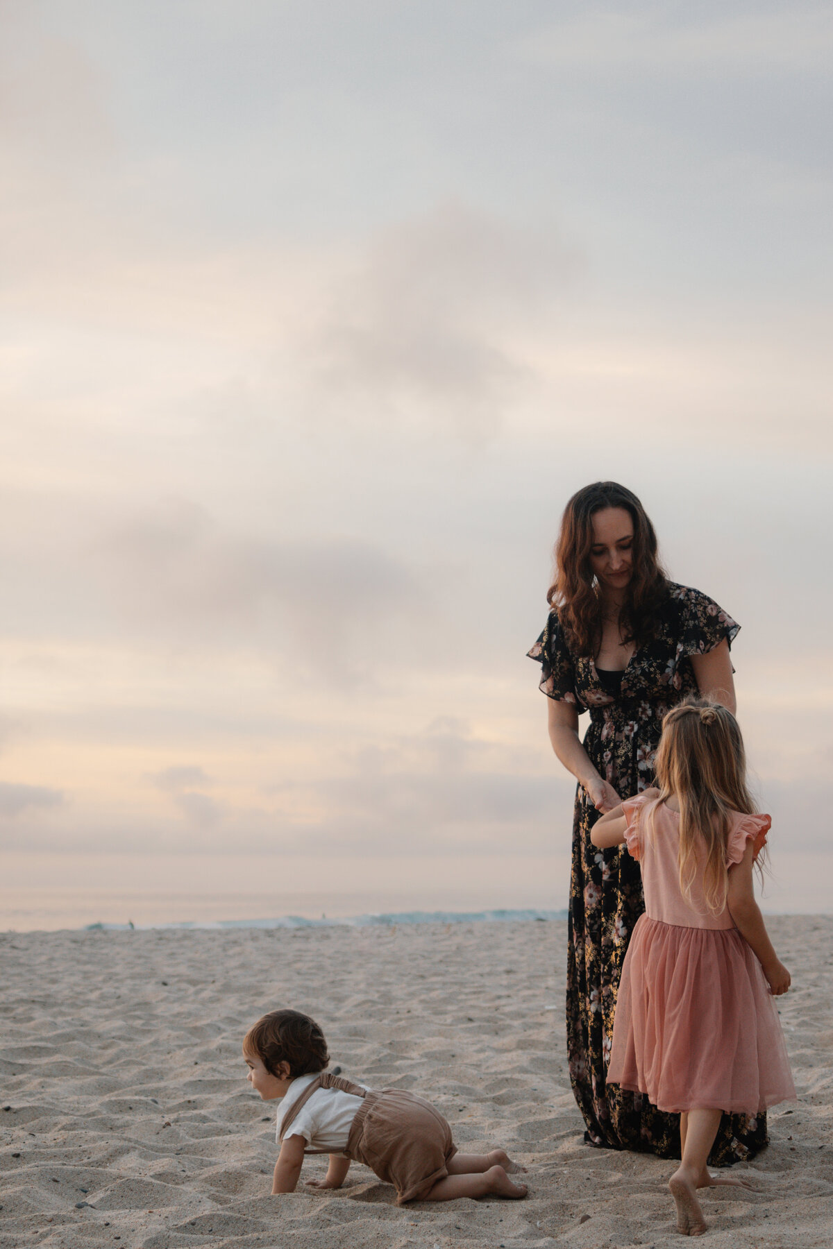 mother candidly hands her daughter a seashell while her infant son crawls on the beach