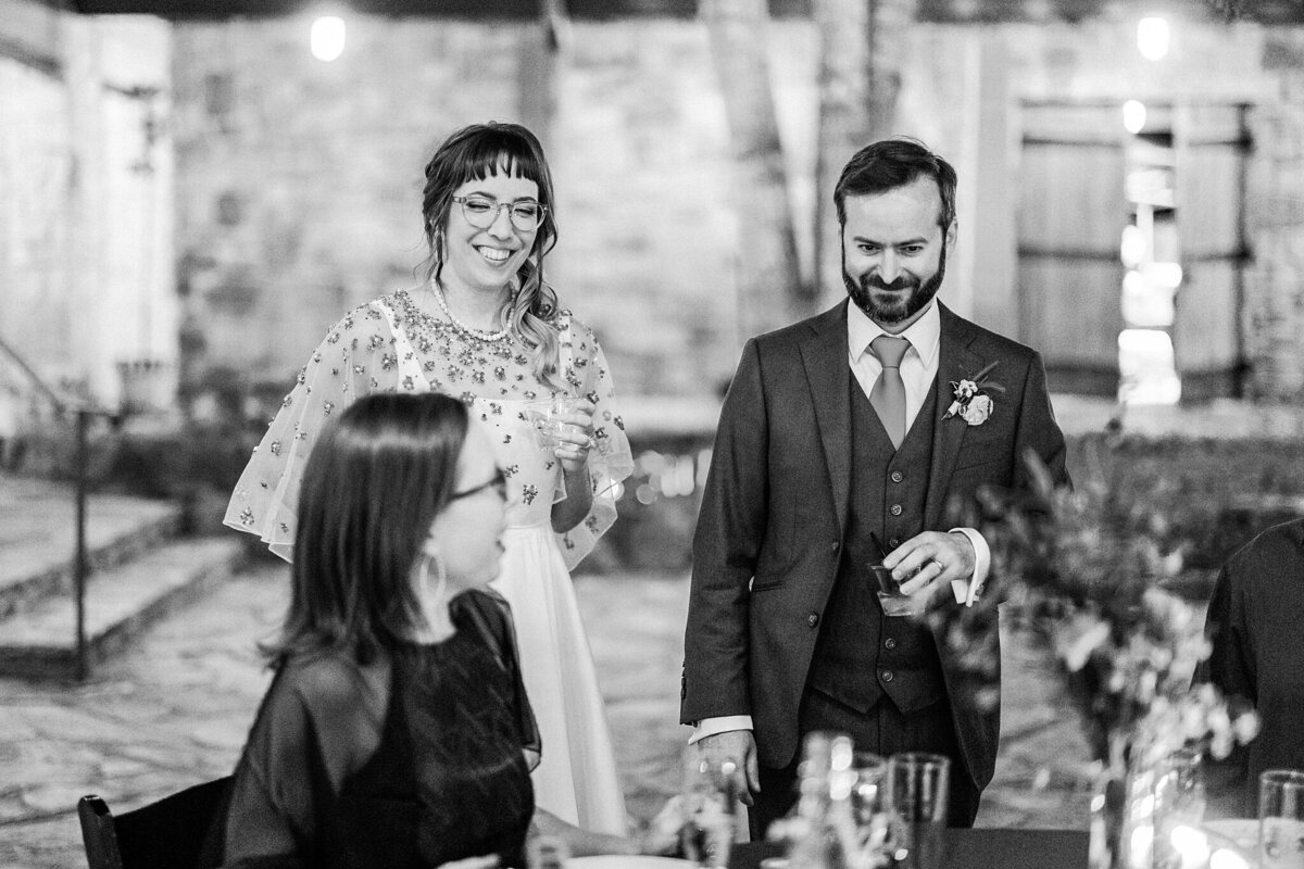 A black and white photo of a bride and groom chatting with a guest at their table during their wedding reception at the Lady Bird Johnson Wildflower Center in Austin, Texas. The bride is on the left and is wearing a white dress with a detailed, mesh shawl. The groom is on the right and is wearing a dark, three piece suit with a boutonniere.