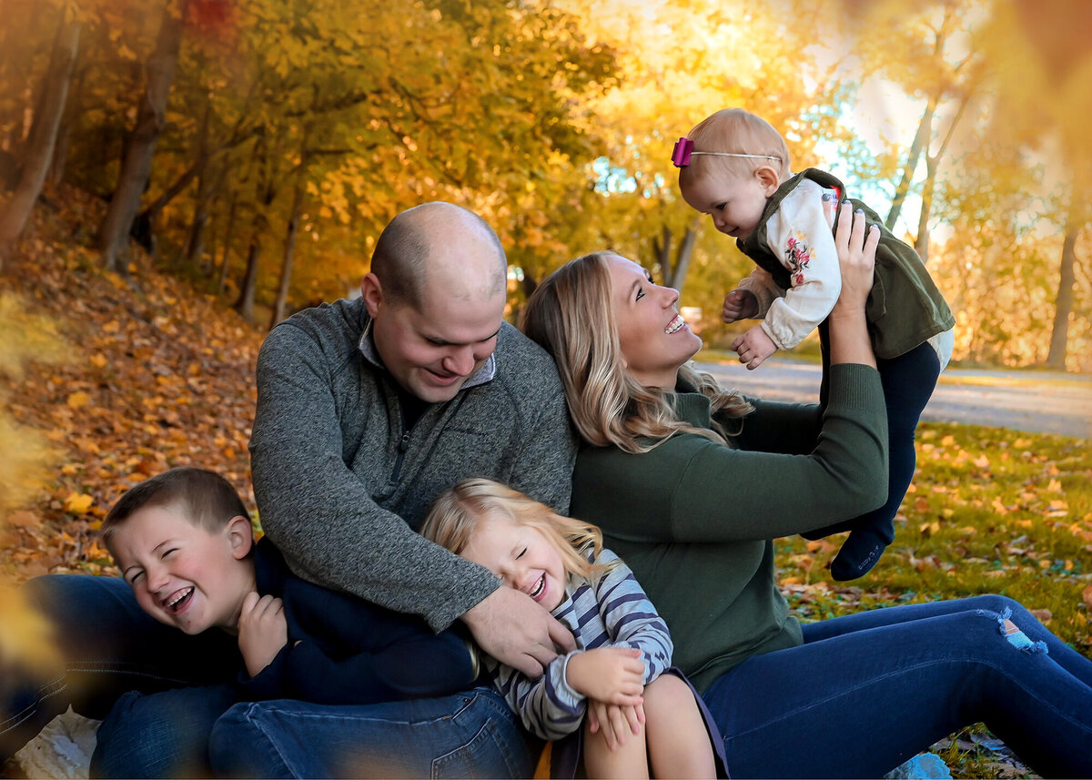 Mom holds baby up and Dad tickles son and daughter in fall foliage