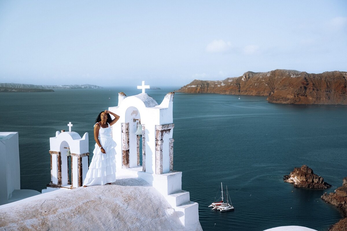 Woman stands on top of building in white dress overlooking the water on a Greek island