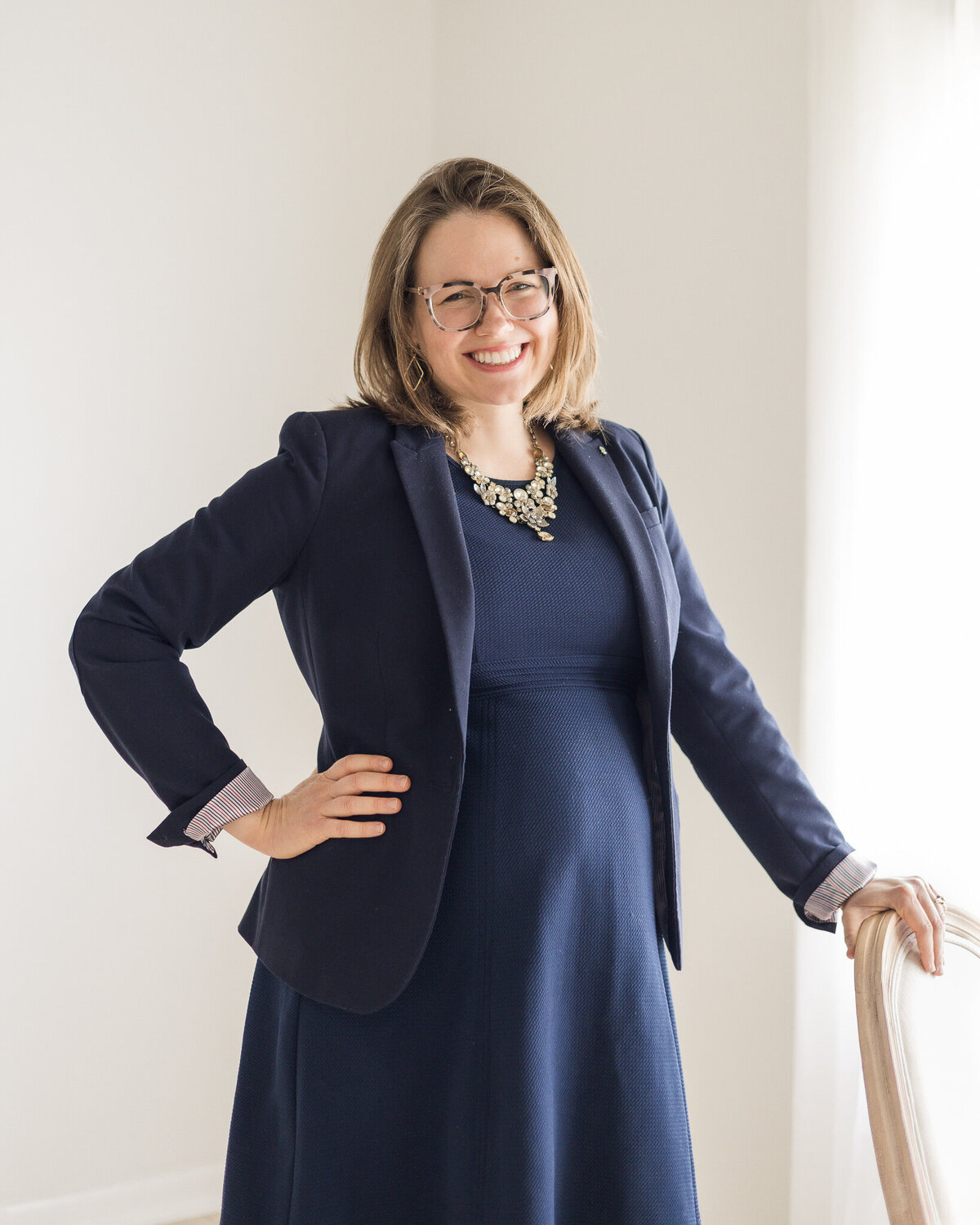 Brunette blonde pregnant woman standing next to a chair with her hand on it, her other hand on her hip, wearing glasses, a navy dress with a navy dress jacket, smiling at the camera