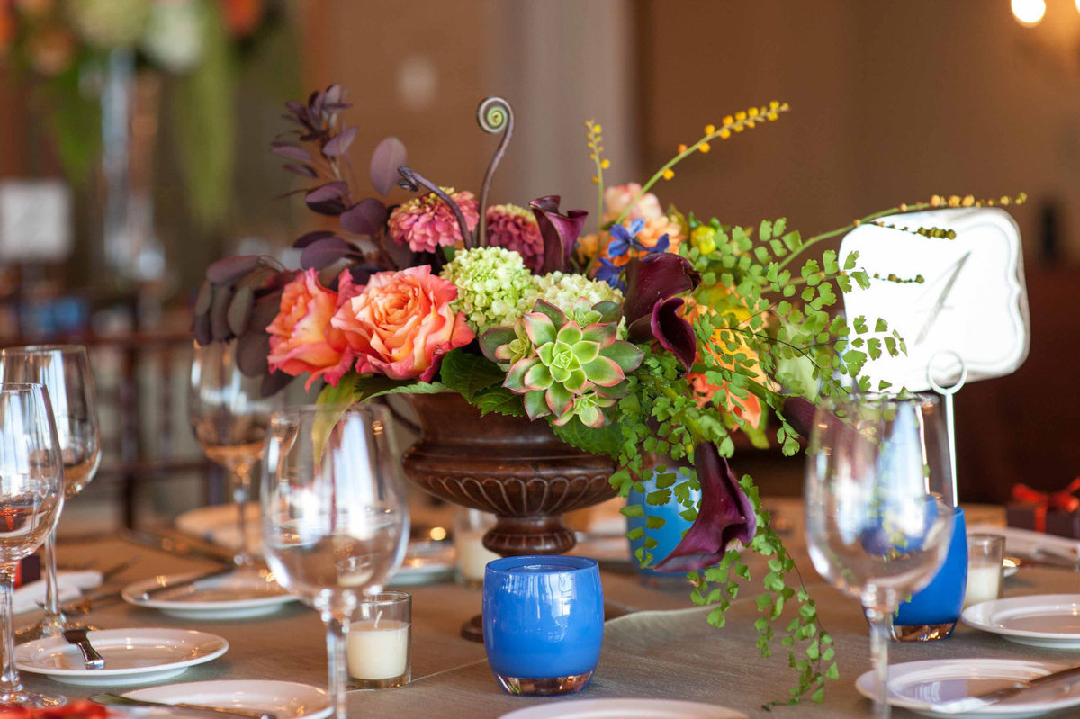 fall centerpiece of orange roses, succulents, purple calla lilies, ferns, green amaranthus, zinnia, fiddlehead fern in brown urn with blue glassybaby candle with matching tall centerpiece in back