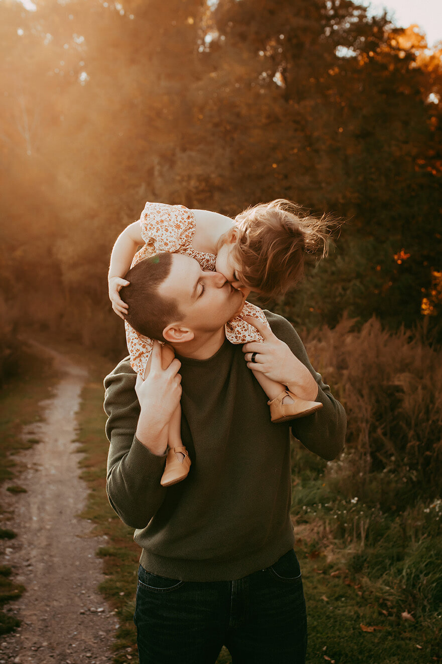 a two year old girl sitting on her dad shoulders kissing at sunset