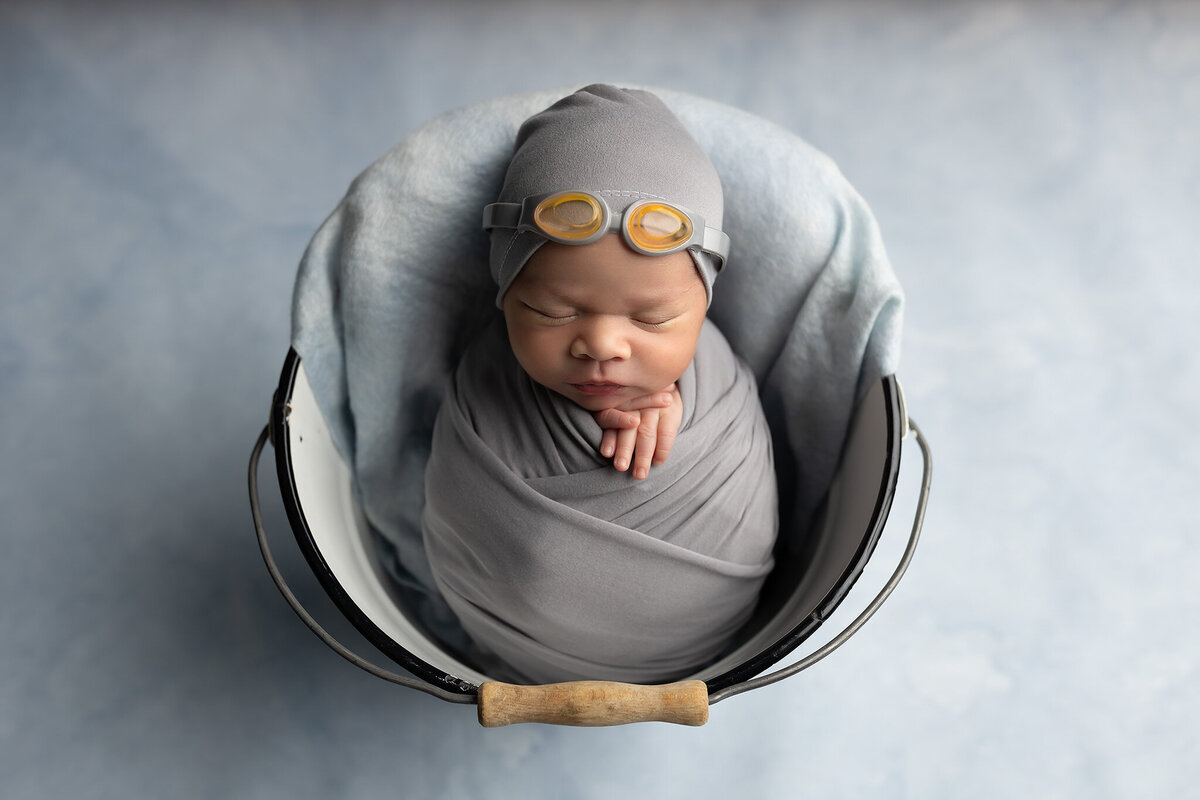 Newborn baby wrapped in gray and wearing swim goggles, posed in a bucket on soft blue backdrop. Dallas baby photographer capturing whimsical newborn photo.