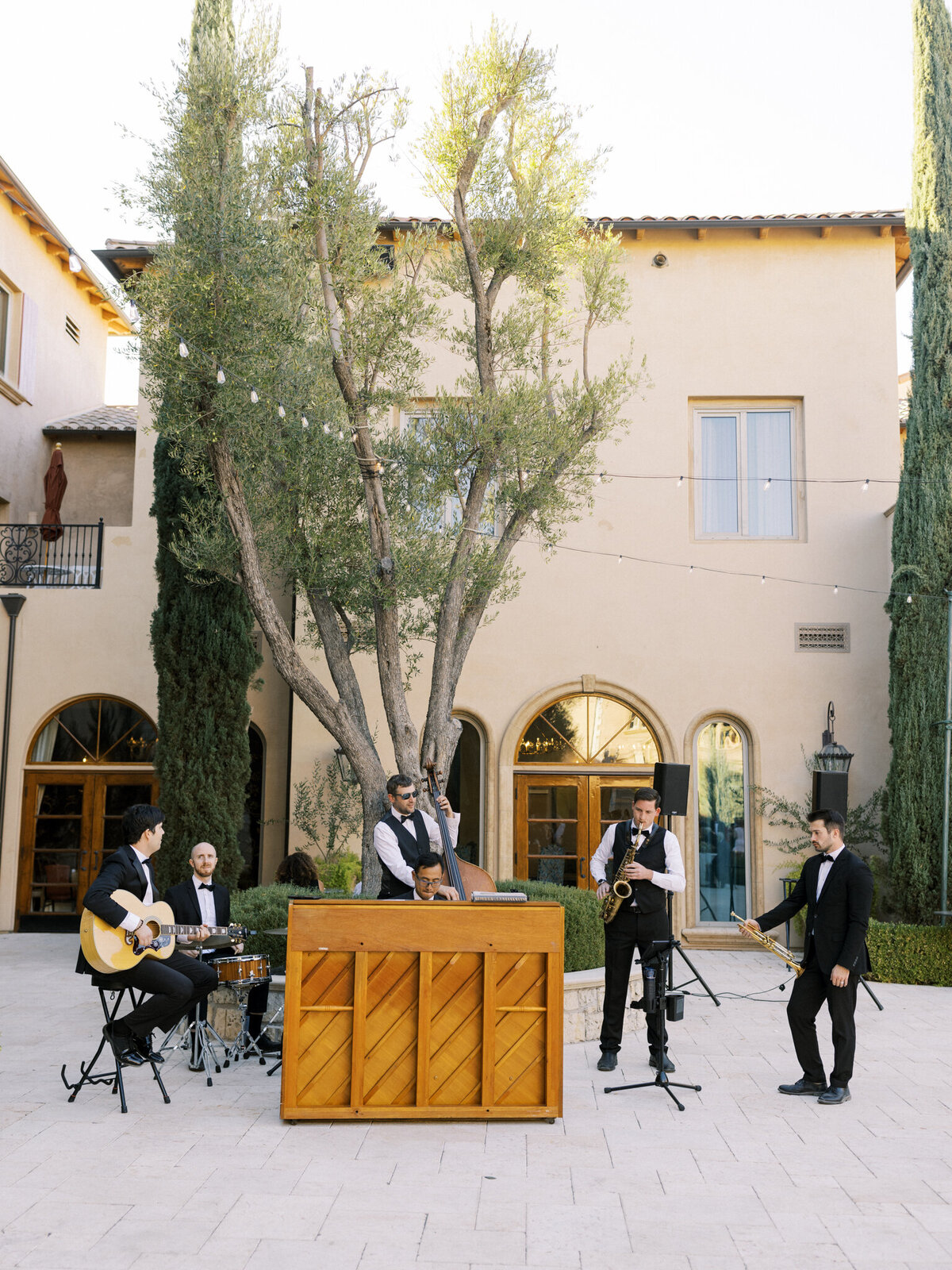  A live band performs outdoors in a courtyard with Mediterranean-style architecture. The group includes a pianist, drummer, guitarist, saxophonist, and double bass player, all dressed in formal black attire. String lights are hung between tall trees and the surrounding beige buildings, creating an elegant and festive atmosphere.