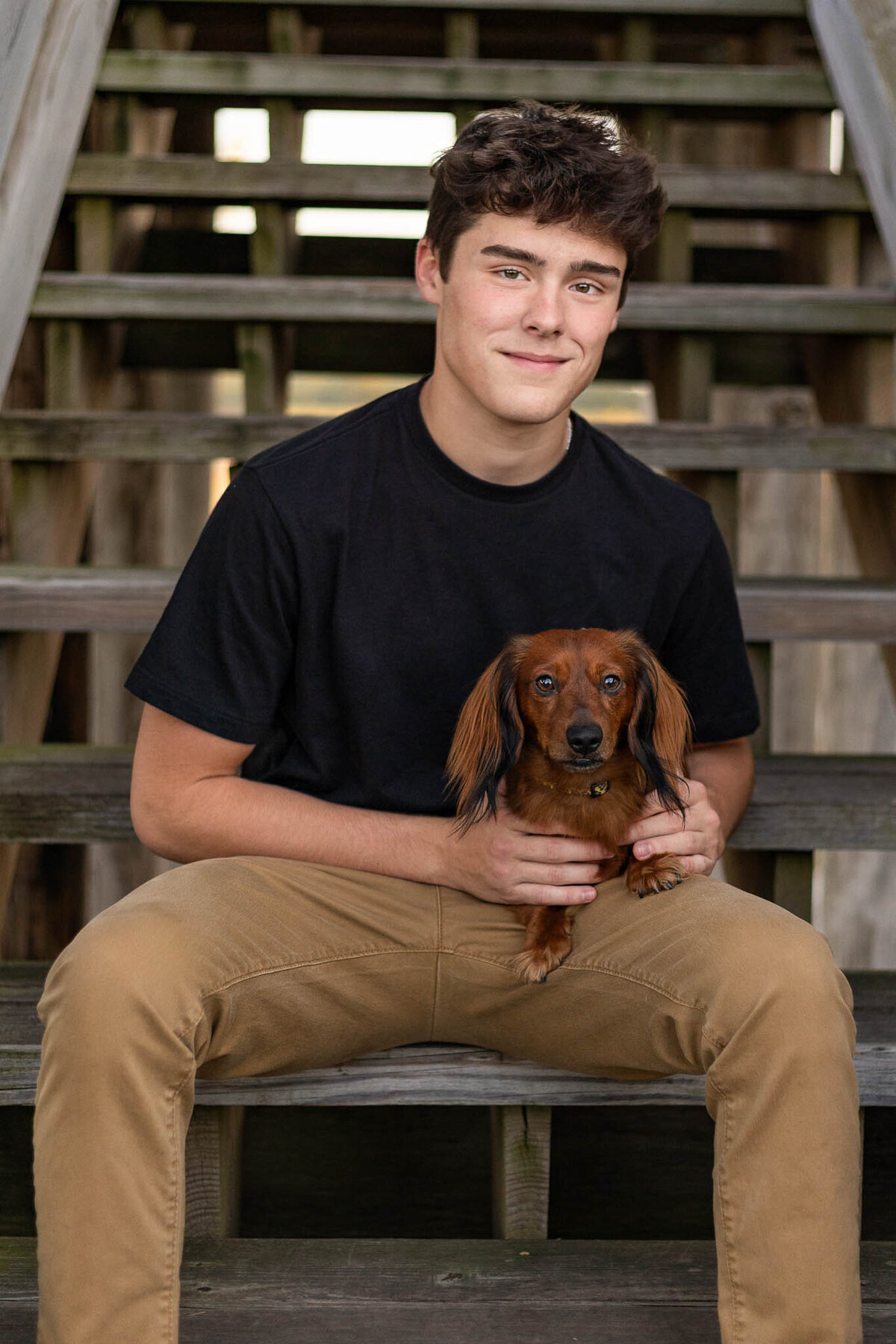 A high school senior holding his brown dachshund dog while sitting on wooden stairs outdoors.