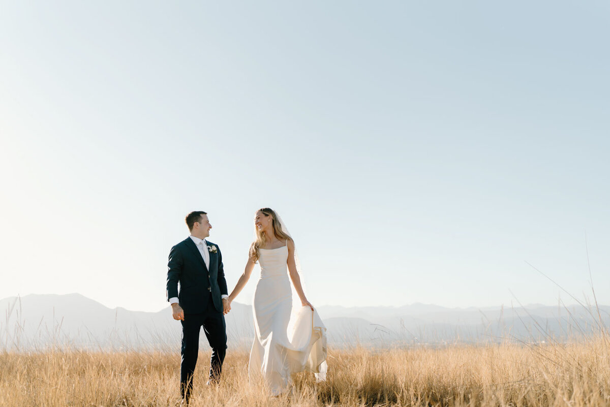 Bride and groom holding hands in a golden field with mountains in the background