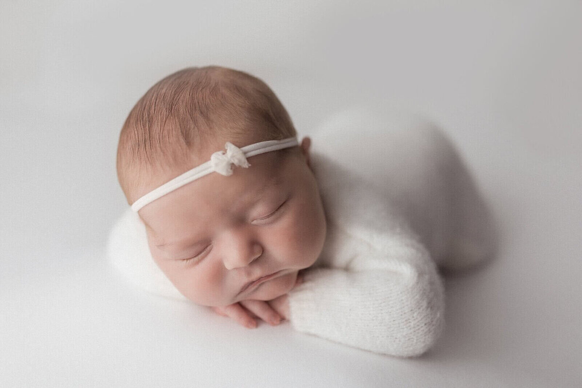 Close-up of a newborn baby girl sleeping peacefully on her tummy, wearing a white knit outfit and a delicate headband with a small bow, set against a soft, white background.