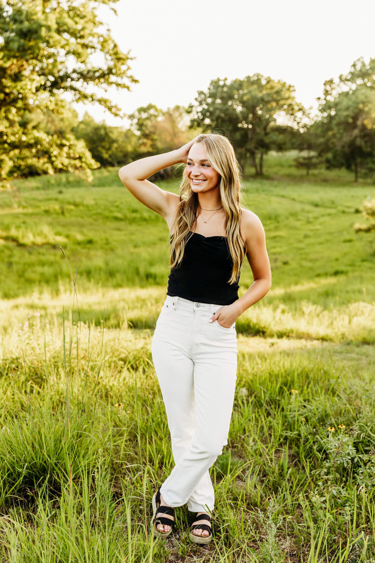 teen girl with brown hair and blonde highlights standing in rolling hills with legs crossed and one hand in hair as she smiles