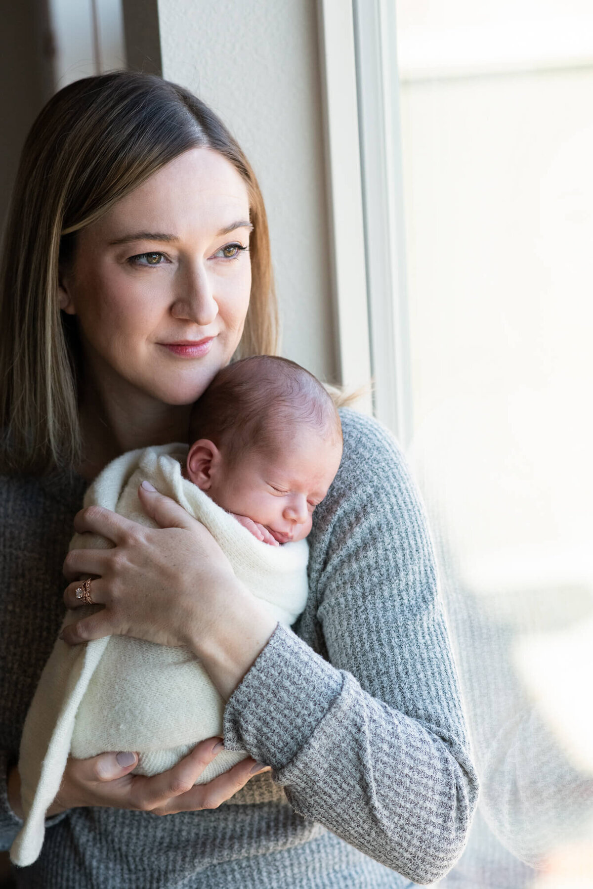 A mother smiles out a window while cradling her newborn baby against her chest for a Denver Newborn Photographer
