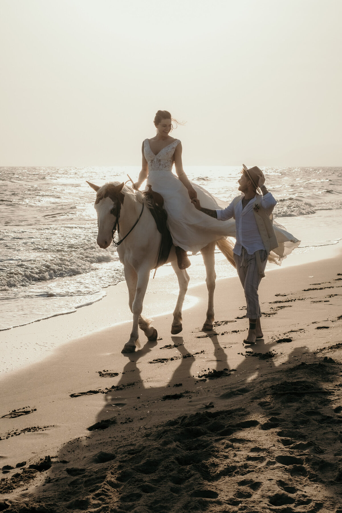 Die Braut reitet auf dem Pferd am Meeresufer. Ihr Bräutigam läuft sie anblickend neben ihr.