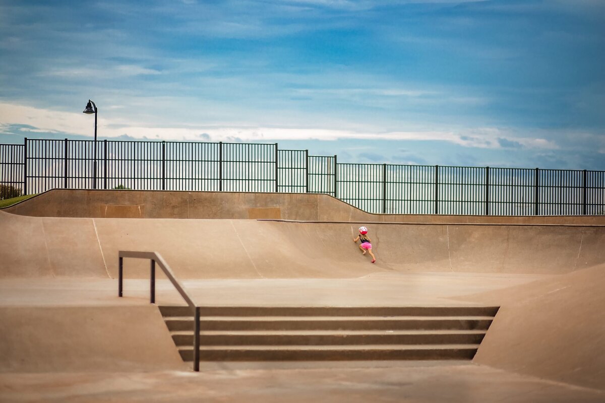 A wideshot of an empty skatepark shows a young toddler in a bicycle helmet trying to climb the ramp.