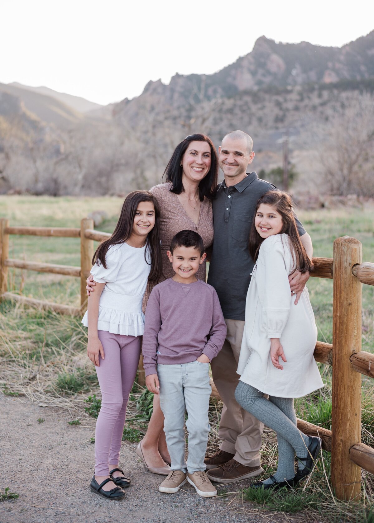 Mom, dad, son, and two girls stand with their arms around each other in front of a wooden fence with the flat irons of Boulder, Colorado behind them captured by denver family photographer