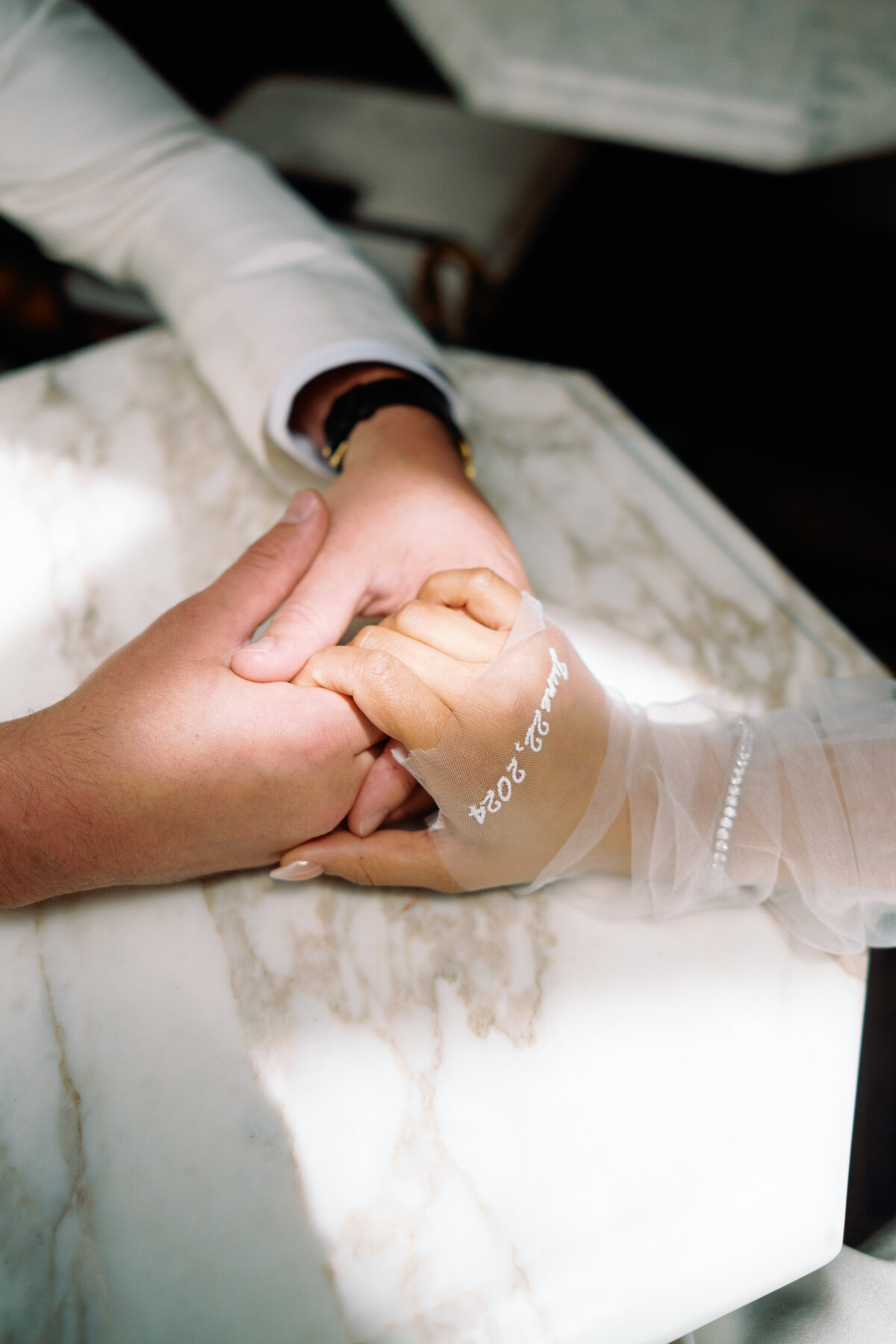 Bride and groom holding hands across a marble table, with custom embroidered wedding gloves reading “Aug 2024,” captured during Japna and Chris’s Hotel Chelsea elopement in New York City by wedding photographer Perry Hancock.