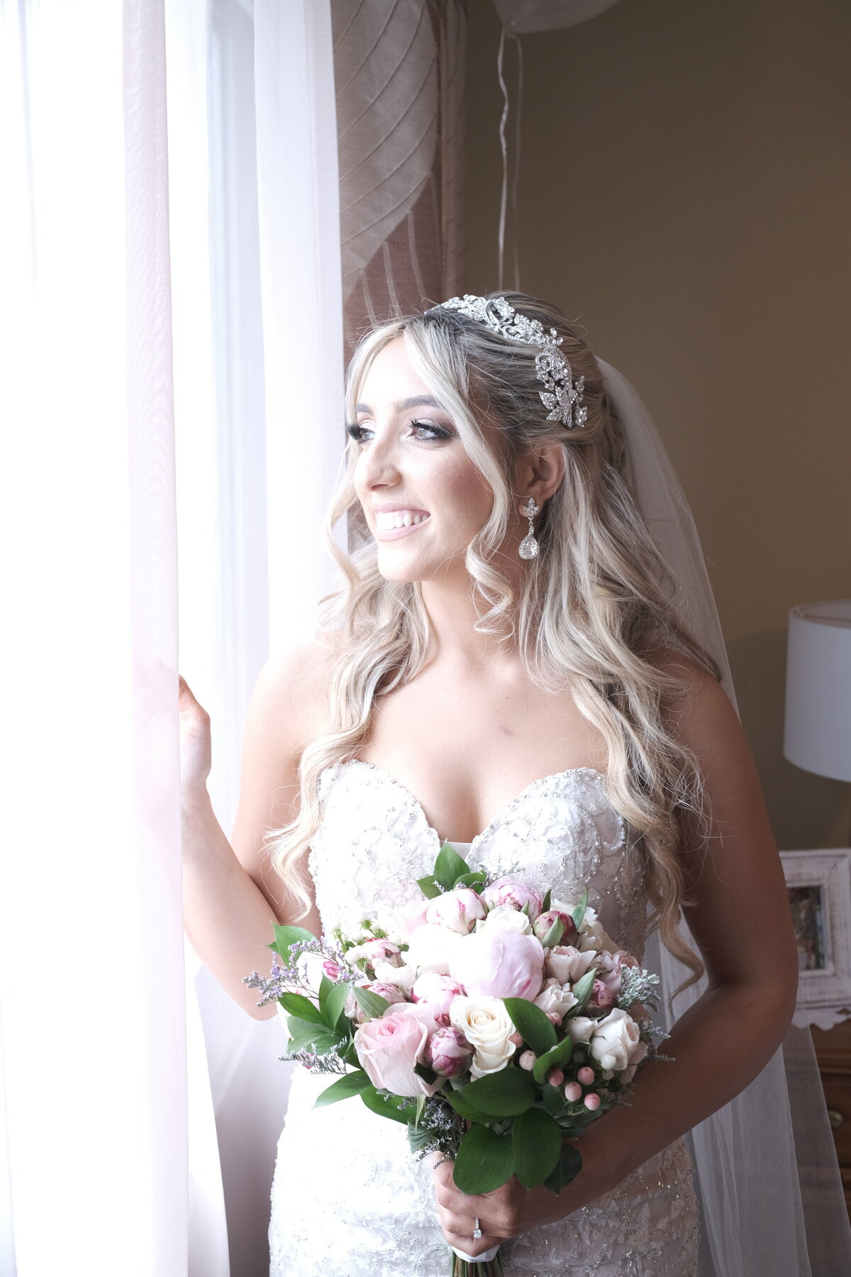 A bride gazes out of a window, her elegant bridal gown beautifully illuminated by natural light. This image captures a serene and reflective moment, highlighting the bride’s grace and the soft, atmospheric setting of her bridal portrait.