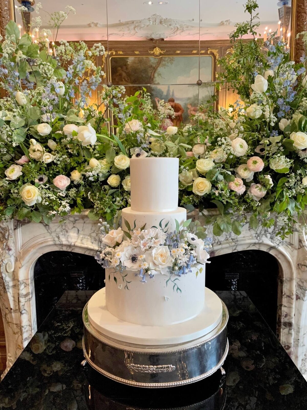 A three tiered cake with lots of sugar flowers in front of a fire place covered in flowers