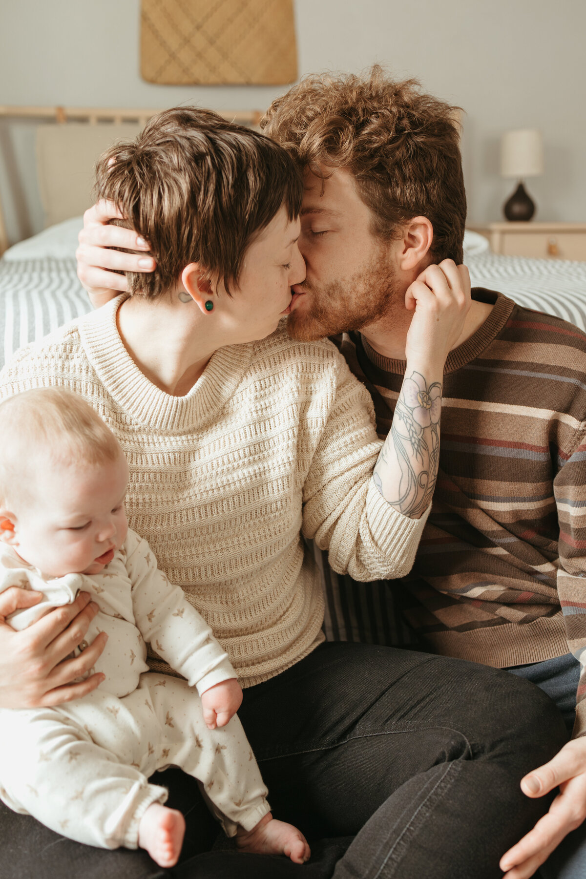 mom and dad holding neck and head while kissing with baby girl in white onesie during in home session in longmont colorado