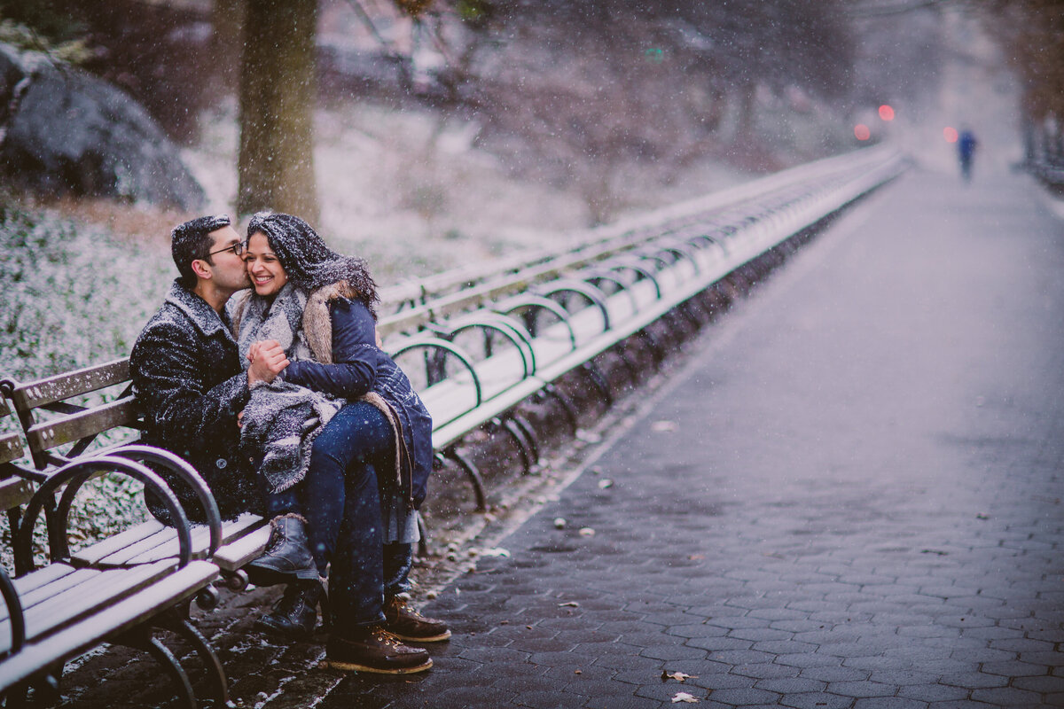 A couple kissing on a bench at the riverside park in new york city during their winter snow storm engagement session