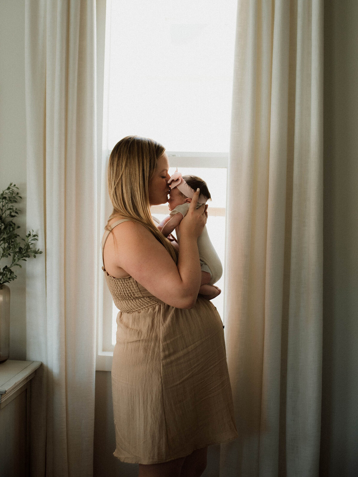 Mom kissing baby tenderly by window light during at-home newborn session in Menifee.