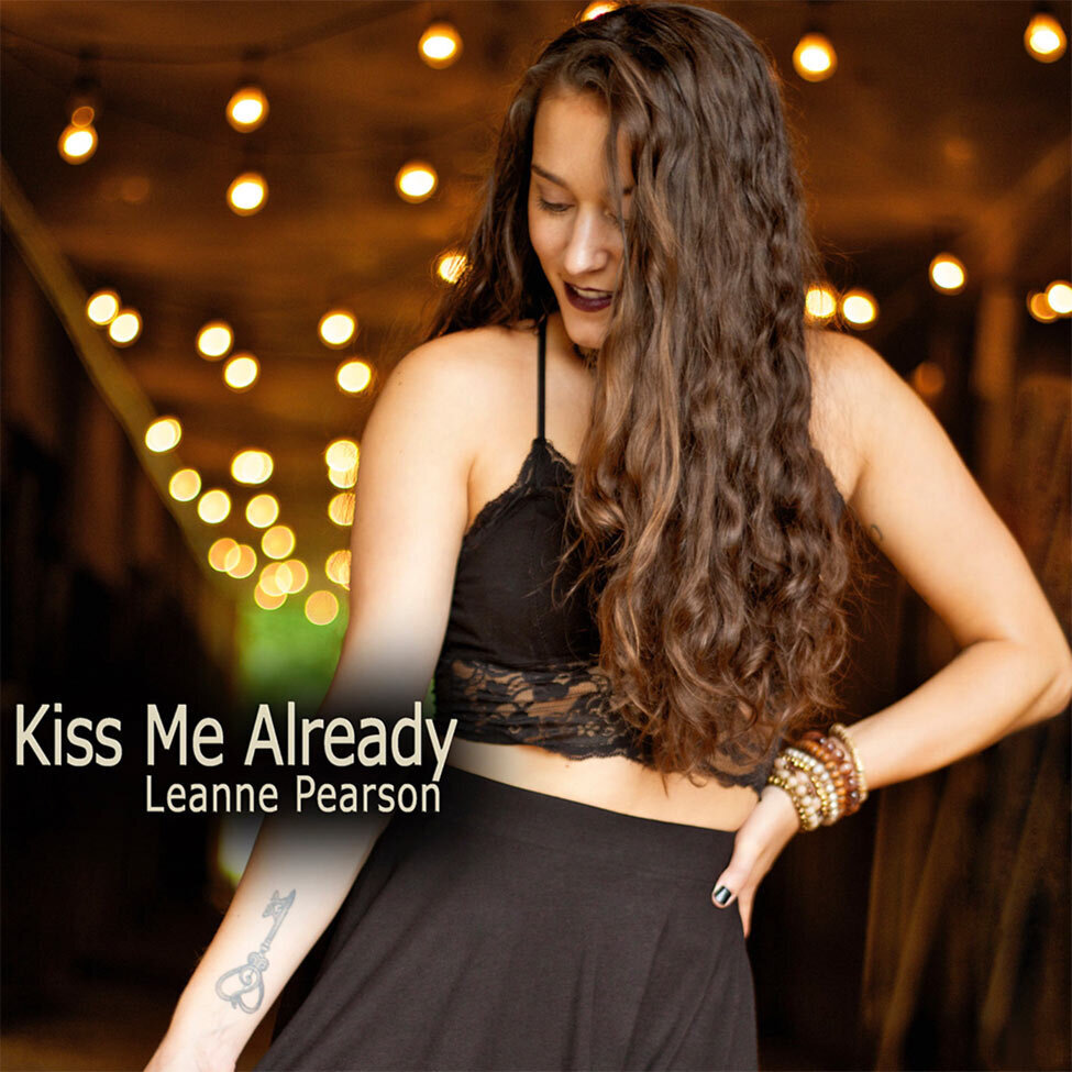 Country Music Single Cover Title Kiss Me Already Artist Leanne Pearson in black top and skirt looking down gold lights in the wood ceiling behind her
