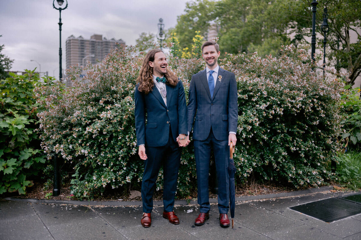 The two grooms are happily chatting, with plants in the background. NYC City Hall Elopement Image by Jenny Fu Studio