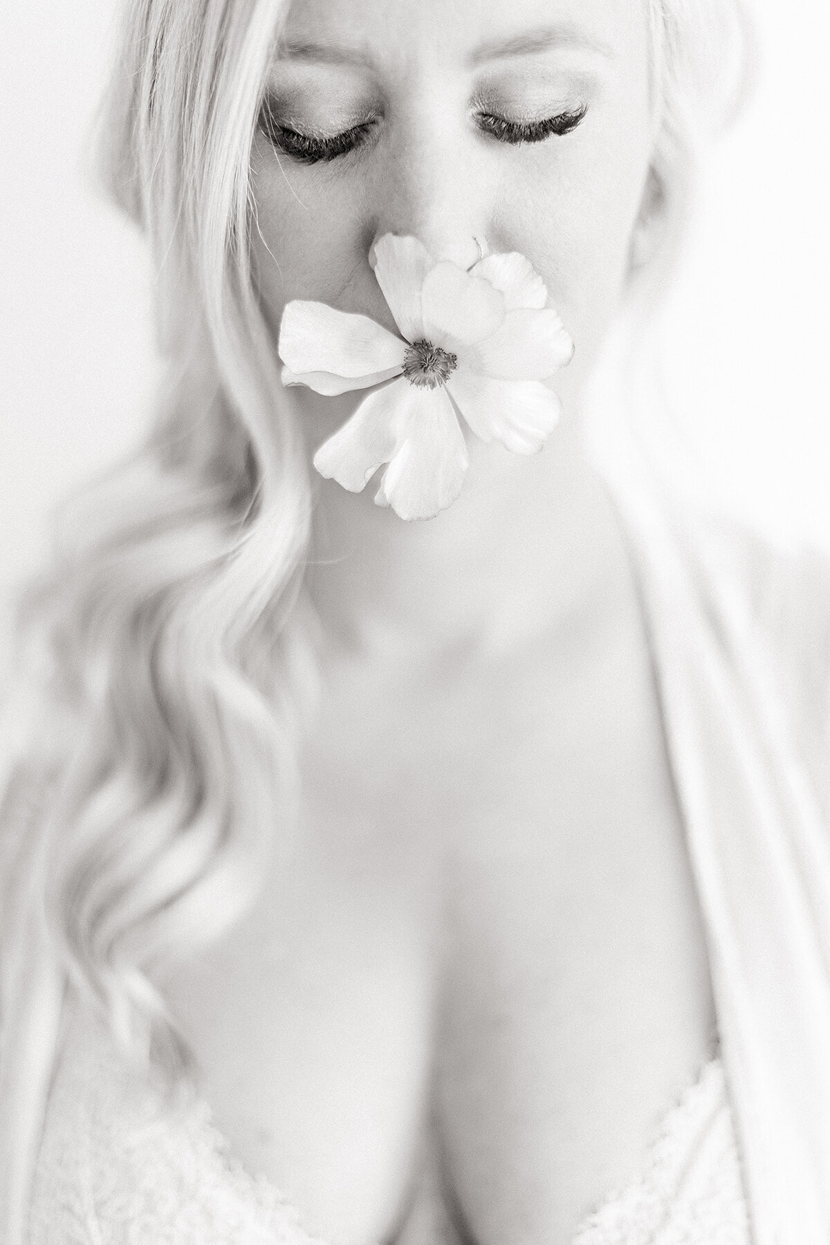 Light filled black and white boudoir photo of a woman wearing lace lingerie while she has a fresh flower in her mouth.