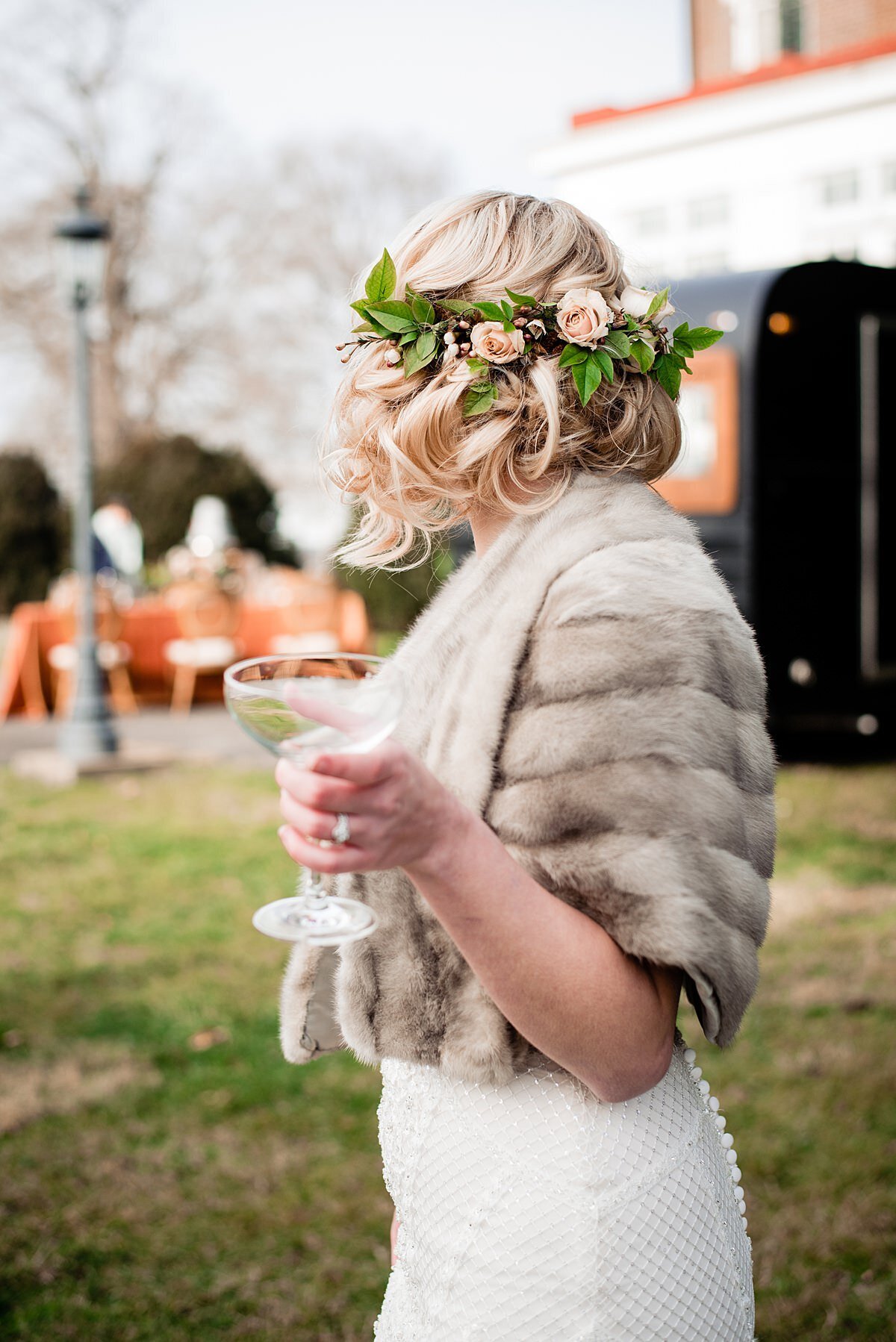 A bride in a lace mermaid dress with buttons down the back wearing a light brown fur wrap is holding a coupe glass of champagne as she looks away from the camera showing off her flower crown of lemon leaves and blush tea roses.