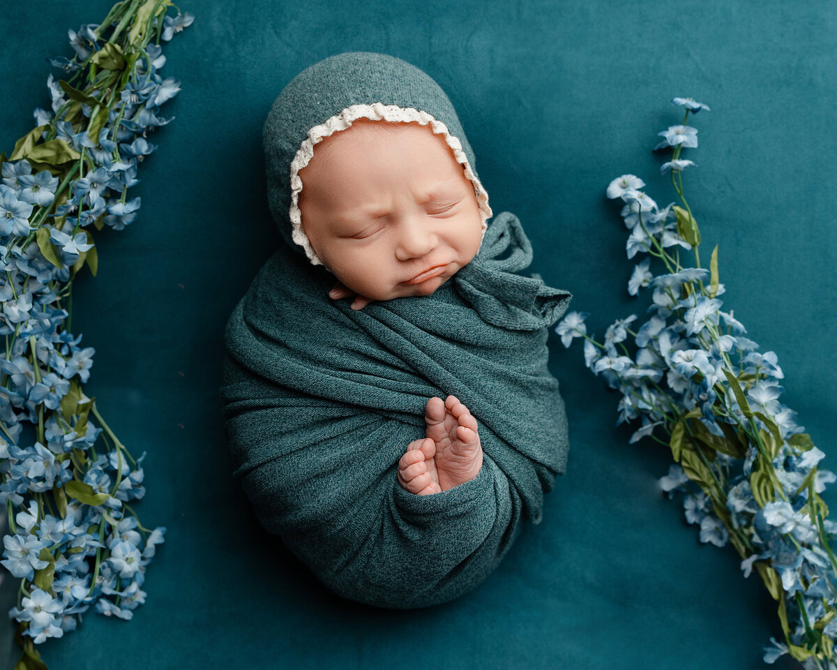 Studio portrait of a newborn baby girl wrapped snugly in teal and accented by floral details, creating a timeless and serene composition. Captured by Inland Empire newborn photographer Kaitlyn Dawn.