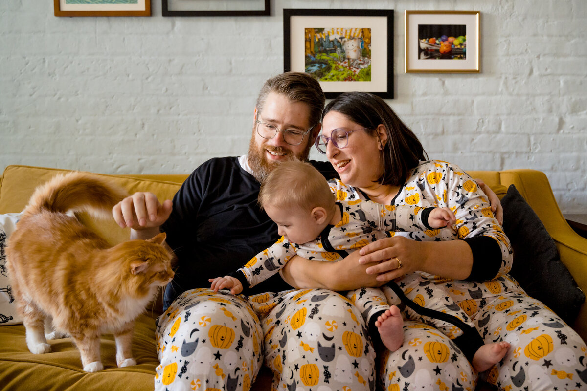 Parents and their baby in matching pajamas sitting on a couch petting a cat.