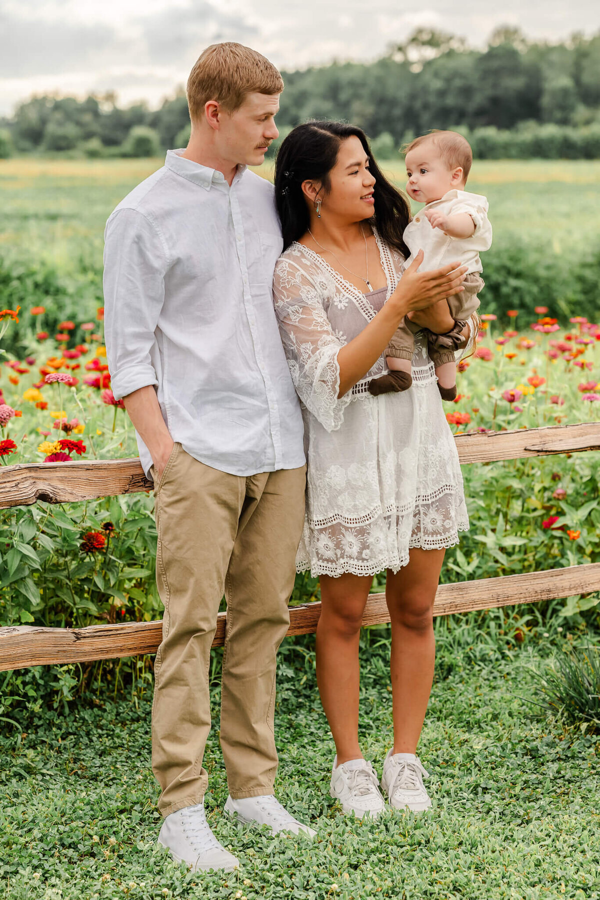 A mother holds her infant son while her husband looks on. They are standing in front of a field of flowers in Chesapeake, Virginia.