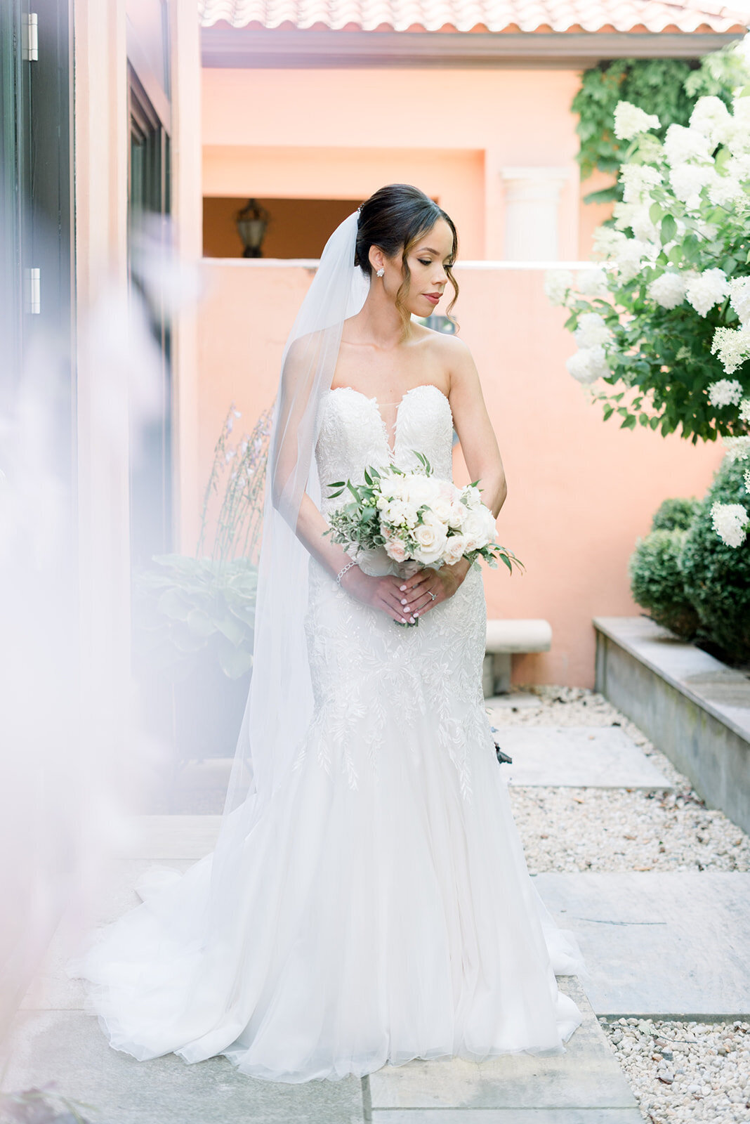 A bride holds a white bouquet while walking through a garden patio in her dress and gazing down her shoulder at her Glenmere Mansion Wedding