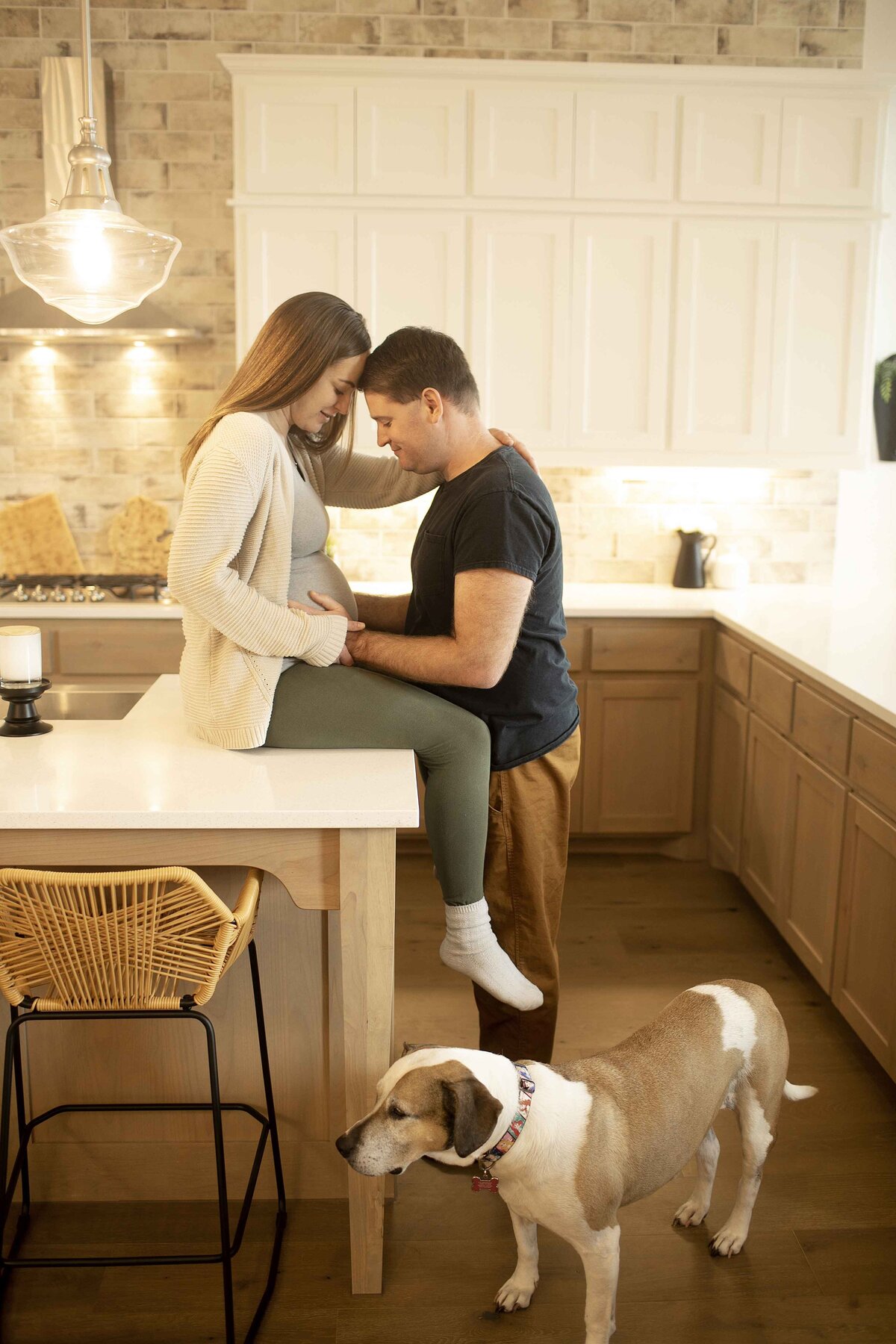 A heartwarming maternity moment of mom in a tank top, off-the-shoulder sweater, and leggings, standing in the kitchen with her husband, both sharing a joyful look while they rest their hands on her belly, captured by Chunky Monkey Photography.