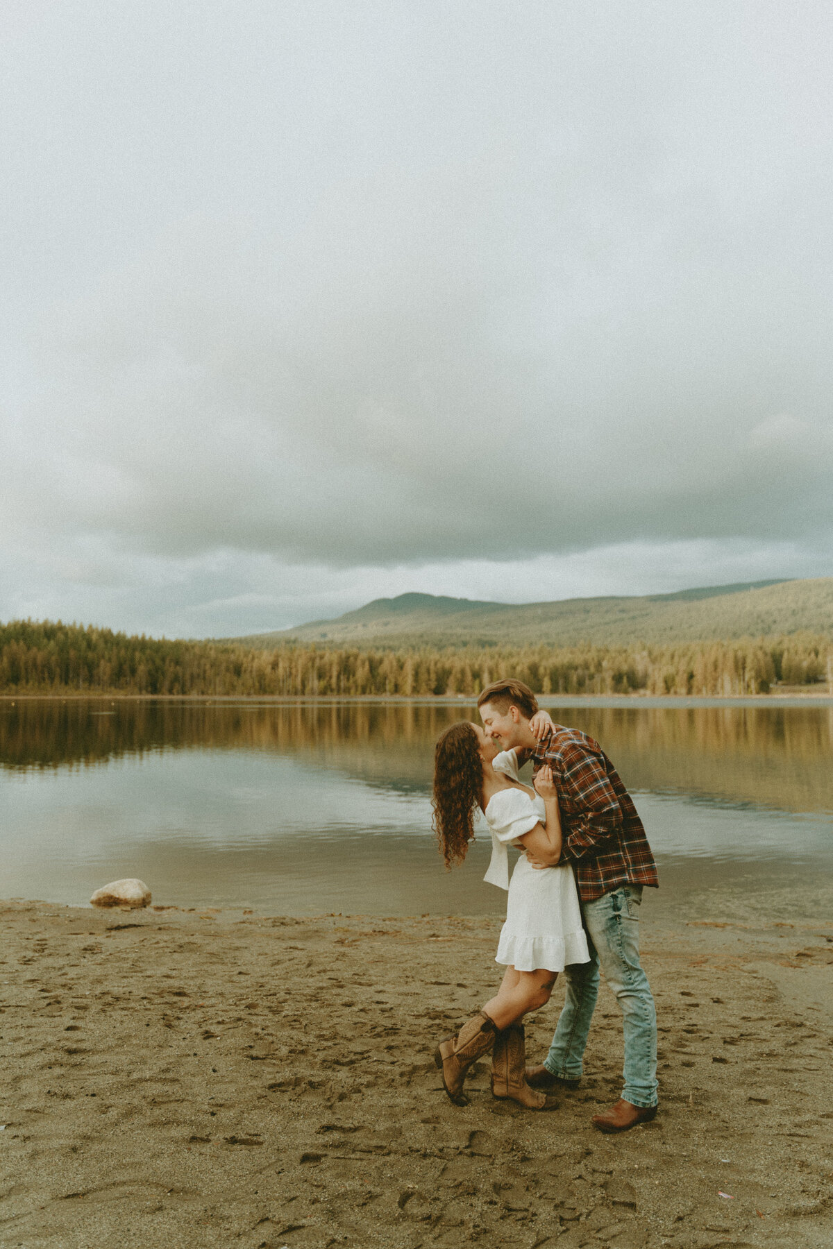 photo of a guy and girl kissing at the lake with mountains in the background