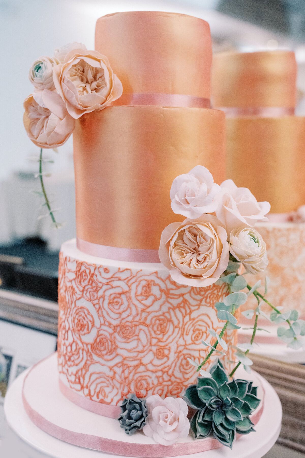 Rose gold 3 tier wedding cake with stenciled roses on bottom tier, sugar flower roses, ranunculus, succulents, and eucalyptus