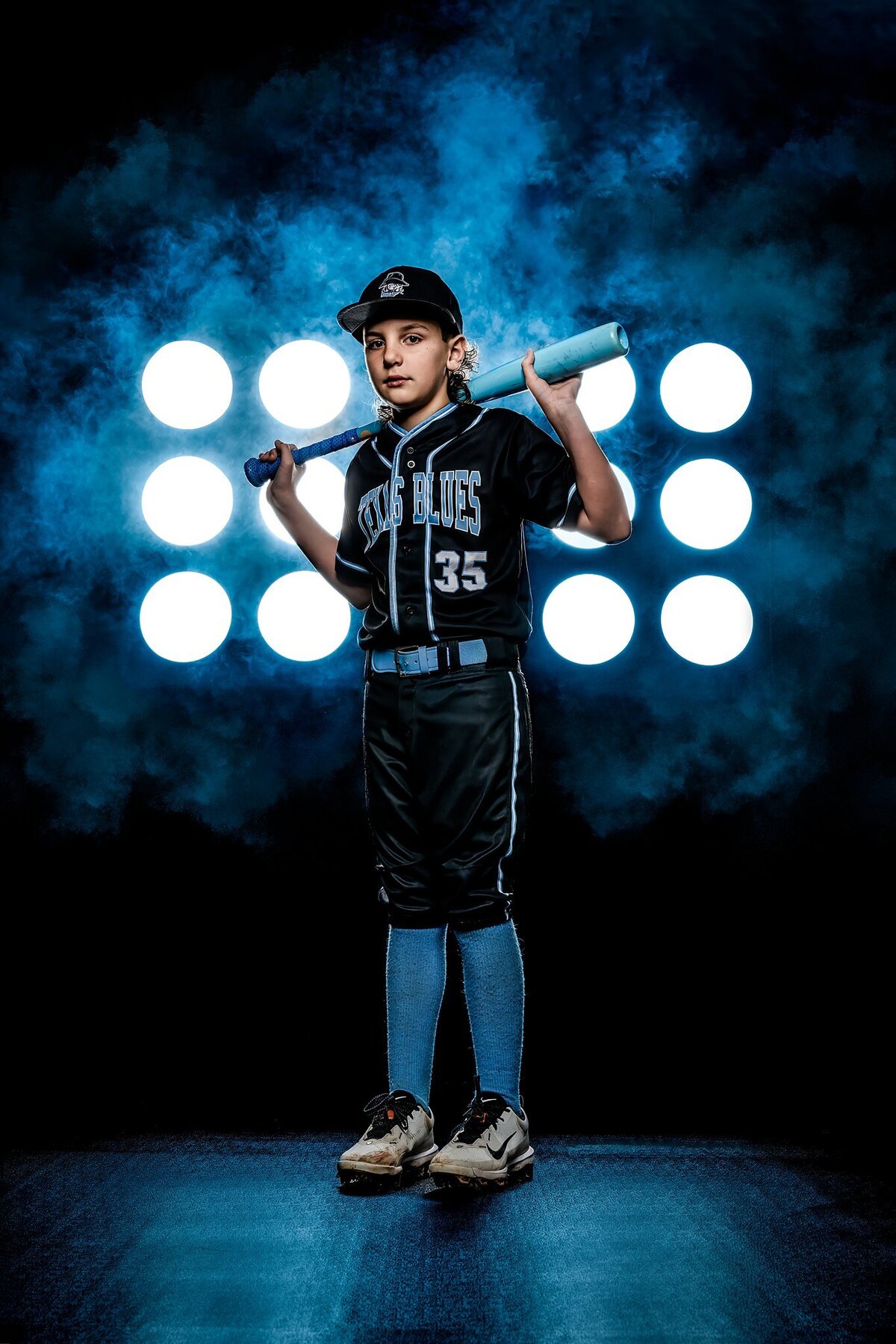 The image captures a young boy proudly wearing his Texas Blues baseball uniform for his annual baseball photoshoot. He stands confidently in a professional studio setting, framed by a striking black backdrop that enhances the overall composition. Two Pro mod stadium lights cast a bright glow on him, creating a dramatic effect while illuminating the blue smoking mist swirling in the air. This mist adds an artistic touch, accentuating the lights.  With a baseball bat resting on his shoulder, the young athlete gazes directly into the camera with determination and focus, embodying the spirit of a dedicated player ready to take on challenges. His uniform features vibrant colors and team insignia, showcasing his pride in representing the Texas Blues. The combination of lighting, mist, and his intense expression conveys a sense of energy and enthusiasm for the sport. This photo not only captures a moment in time but also reflects the passion and commitment that youth sports inspire in young athletes.