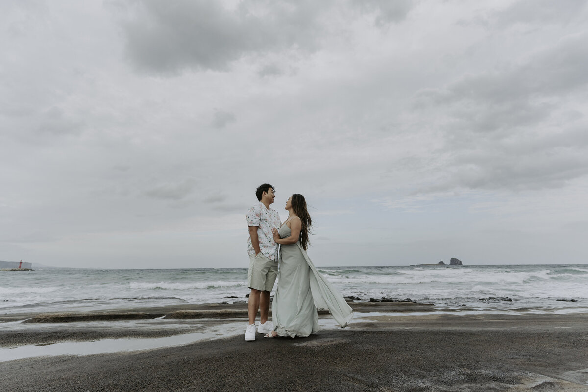 the couple standing at the beach of Jeju island and the bride wears a casual long dress while the groom wears a polo and a shorts