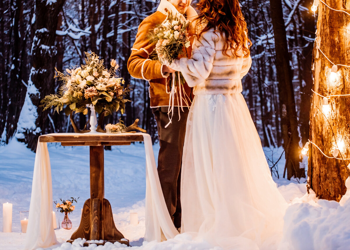 A wedding set up is captured in the snowy forest, with flowers and festoon lighting. The bride wears a fur jacket.
