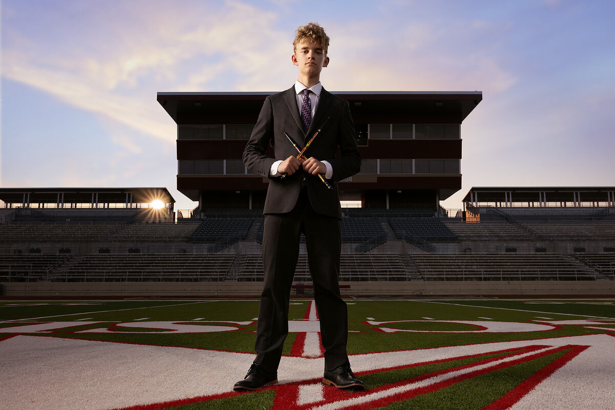 A high school senior stands tall on the football field at Mustang Panther Stadium, holding drumsticks as he strikes a confident pose. The stadium's stands loom in the background, captured during sunset in this Dallas senior photography session.