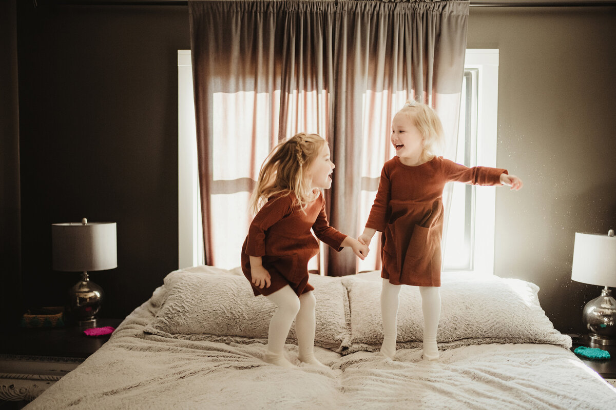 Two little girls are jumping on the bed while looking at one another and laughing and holding hands.