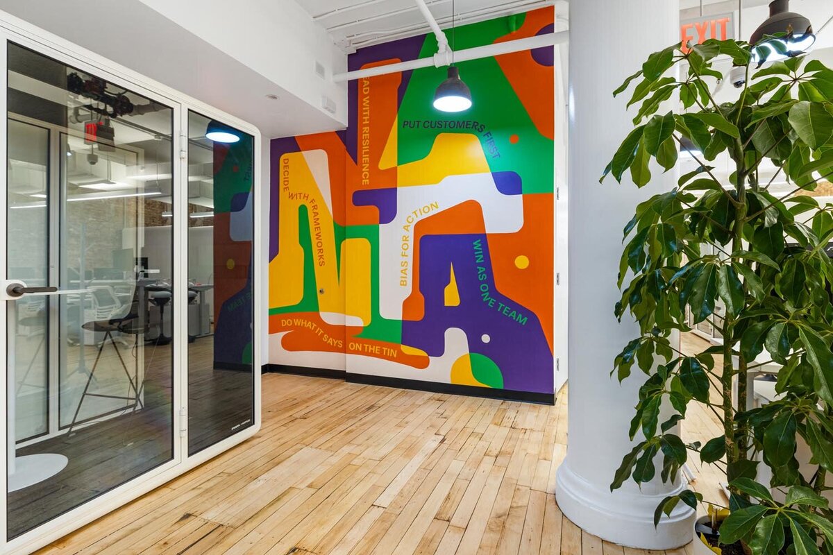 Graphic mural corner with brand values overlaid on bold shapes, next to glass meeting booths and plants.