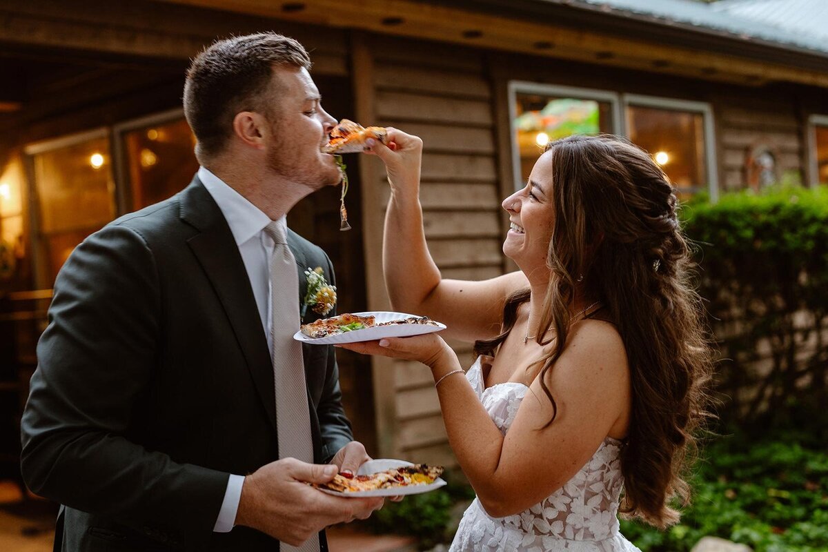 A bride feeds her groom a piece of pizza during a pizza cut at their wedding. 
