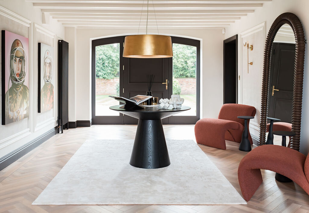 A modern foyer features a black table with decor, two red chairs, a large mirror, wall art, and a gold pendant light on a white rug set against double doors and light wood flooring.